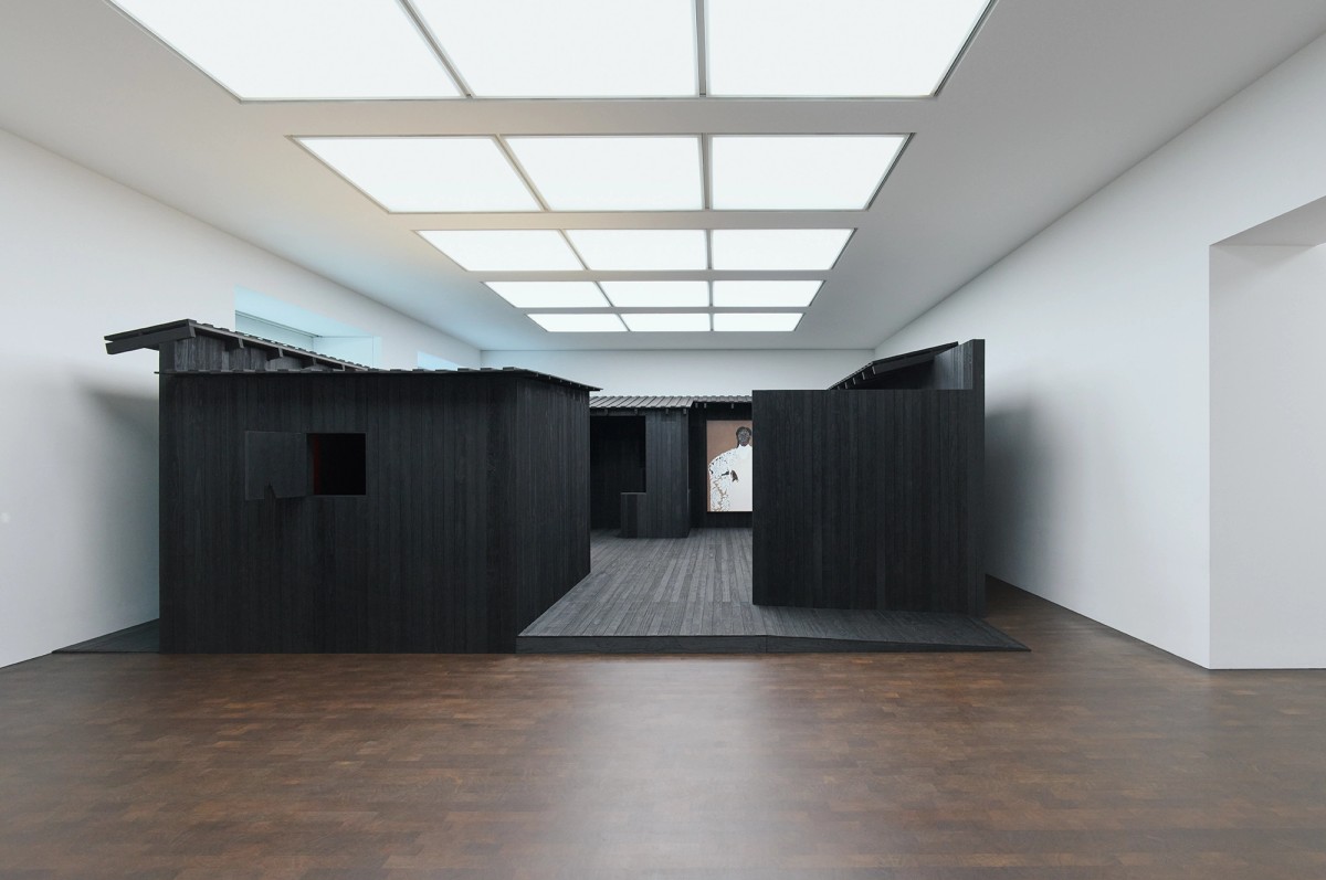 Matte black structures open up into a enterable space, surrounded by white walls and brown flooring. A painting hangs at the back of the structure.