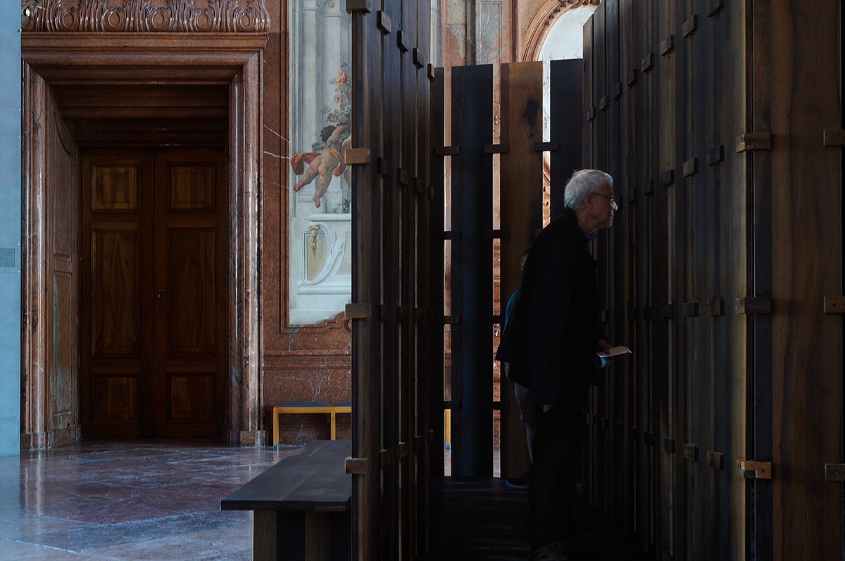 A figure stands inside a wodden structure, with a doorway off to the left and blue painted walls.