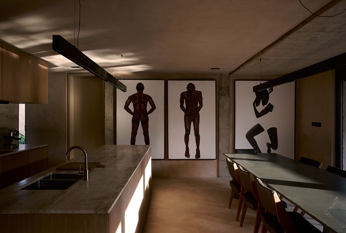 Warmly lit interior featuring a minimalist kitchen and dining area with a marble island, dark dining table, and wood cabinetry. The space is defined by three striking large-scale artworks depicting stylized human figures, hung against a raw concrete wall, creating a dramatic contrast in the refined, modern setting.