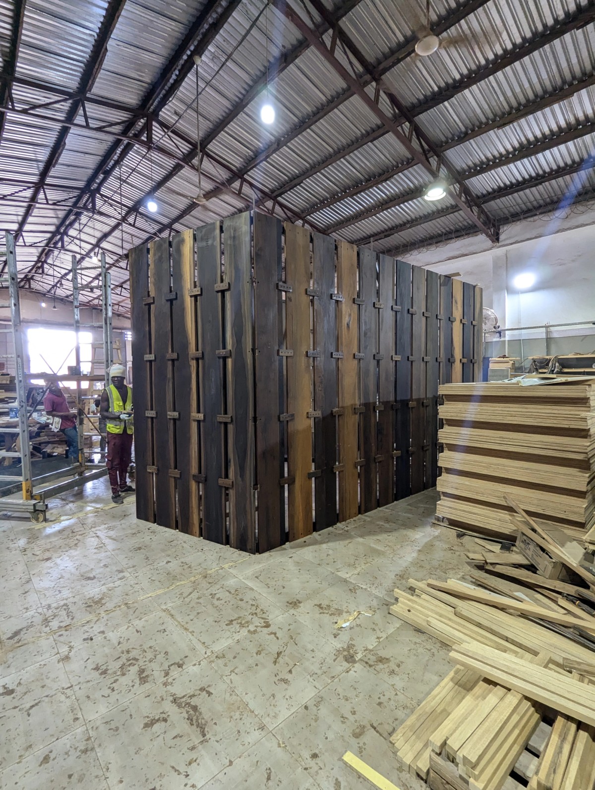Pallets of wood sit opposite a wooden structure, with a worker in a hard hat on the left