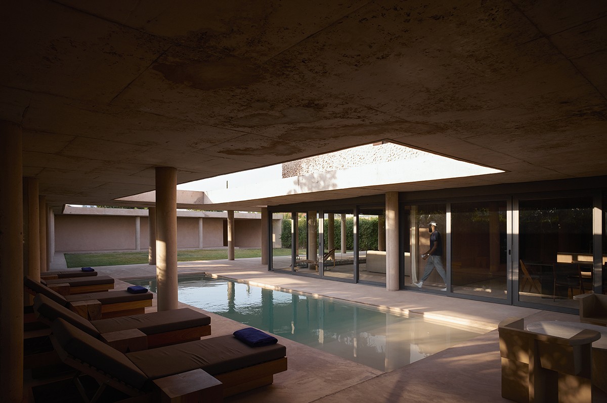 Covered courtyard space with a central swimming pool, surrounded by lounge chairs, concrete columns, and floor-to-ceiling glass doors leading into the house. A large rectangular skylight above allows natural light to pour in, casting shadows across the minimalist architecture and open layout.
