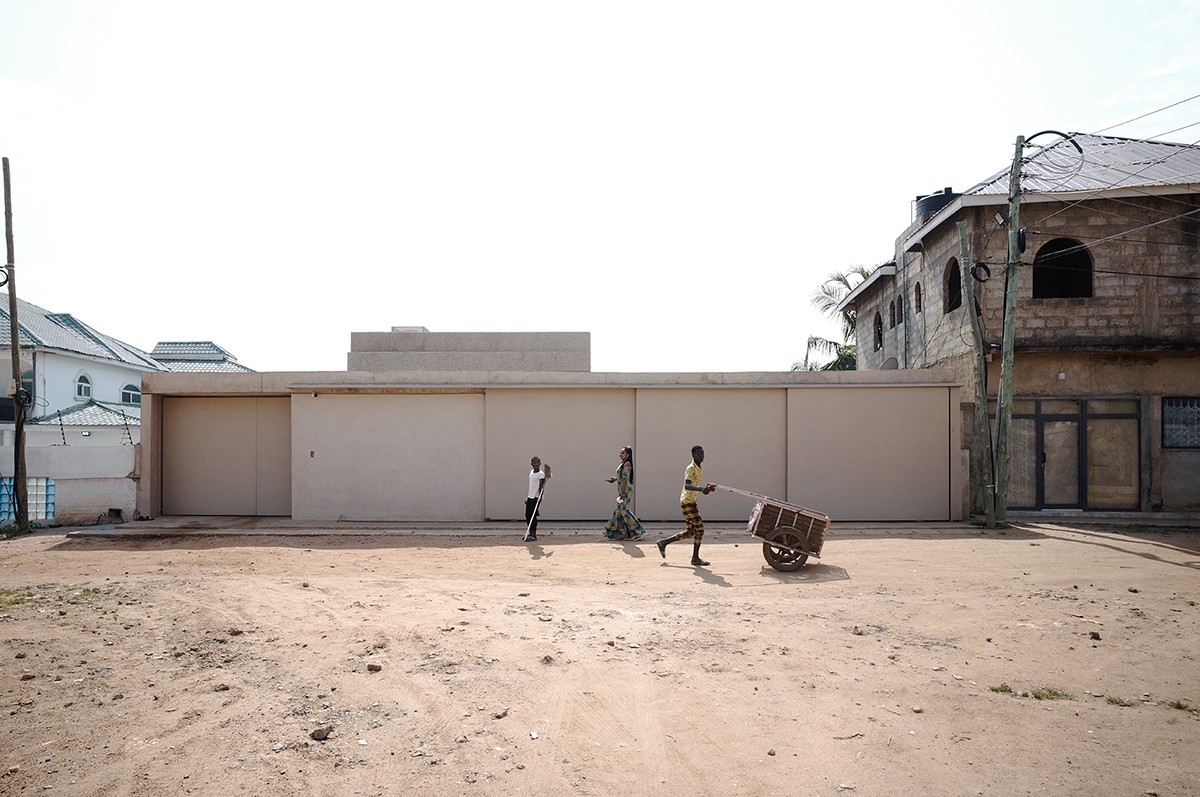 Three figures move in front of a beige structure standing behind sandy ground.