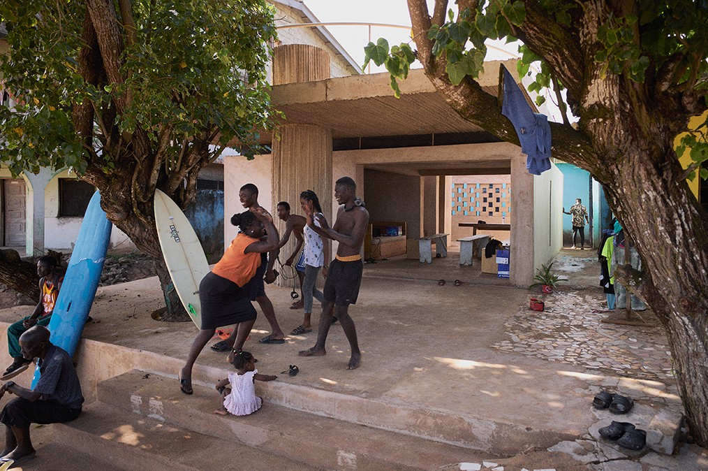 Under the shade of large trees near a beachside building, a group of people interact playfully, with surfboards leaning against the tree trunks and children nearby. The relaxed, open-air space, featuring concrete architecture and colorful walls, creates a vibrant, communal atmosphere filled with everyday activity.