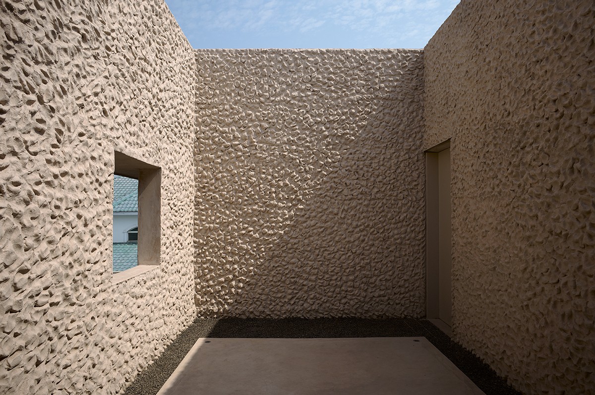 Enclosed courtyard with tall textured walls made of rough, pebble-like plaster, casting sharp shadows in the sunlight. A small square window and a narrow doorway break the surface, offering minimal views to the outside and adding contrast to the stark, sculptural space.