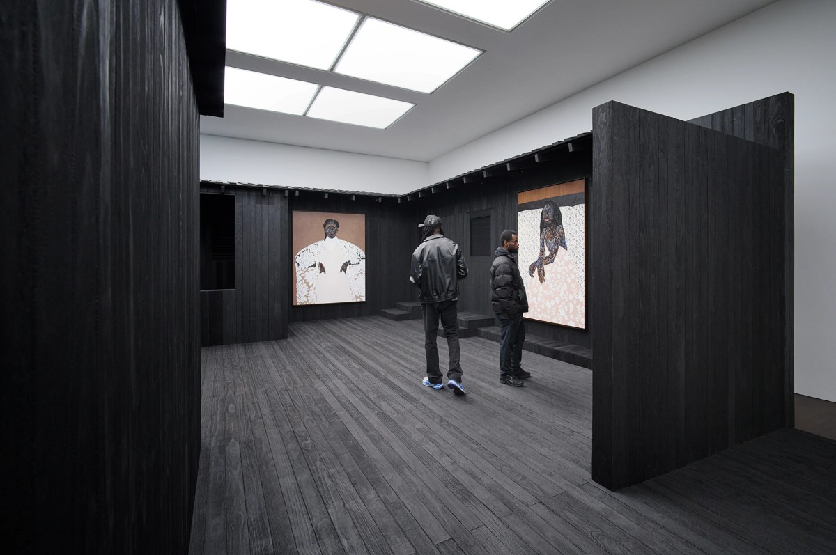 Two people stand in a dark, modern gallery space with black wooden walls and floors, viewing large portrait paintings displayed on the walls under bright ceiling lights.