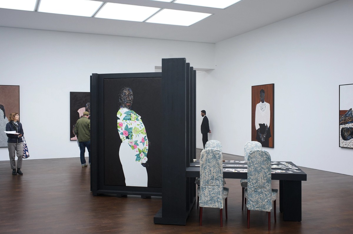 A modern art gallery with visitors, featuring large portraits of figures in patterned clothing on white walls and freestanding panels; a table with two patterned chairs is in the center.