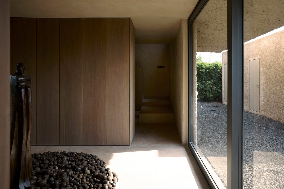 Minimalist interior hallway with warm-toned walls, wood paneling, and a modern sculpture standing on a bed of dark river stones. A large floor-to-ceiling glass window reveals an enclosed outdoor gravel courtyard with a door and lush greenery along the perimeter.