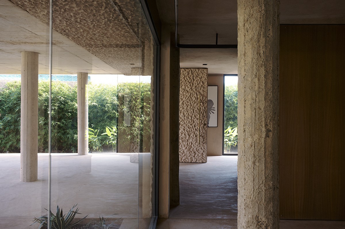 Interior view of a modern home featuring rough-textured columns, smooth concrete floors, and glass walls that open to a lush garden courtyard lined with tall bamboo. 