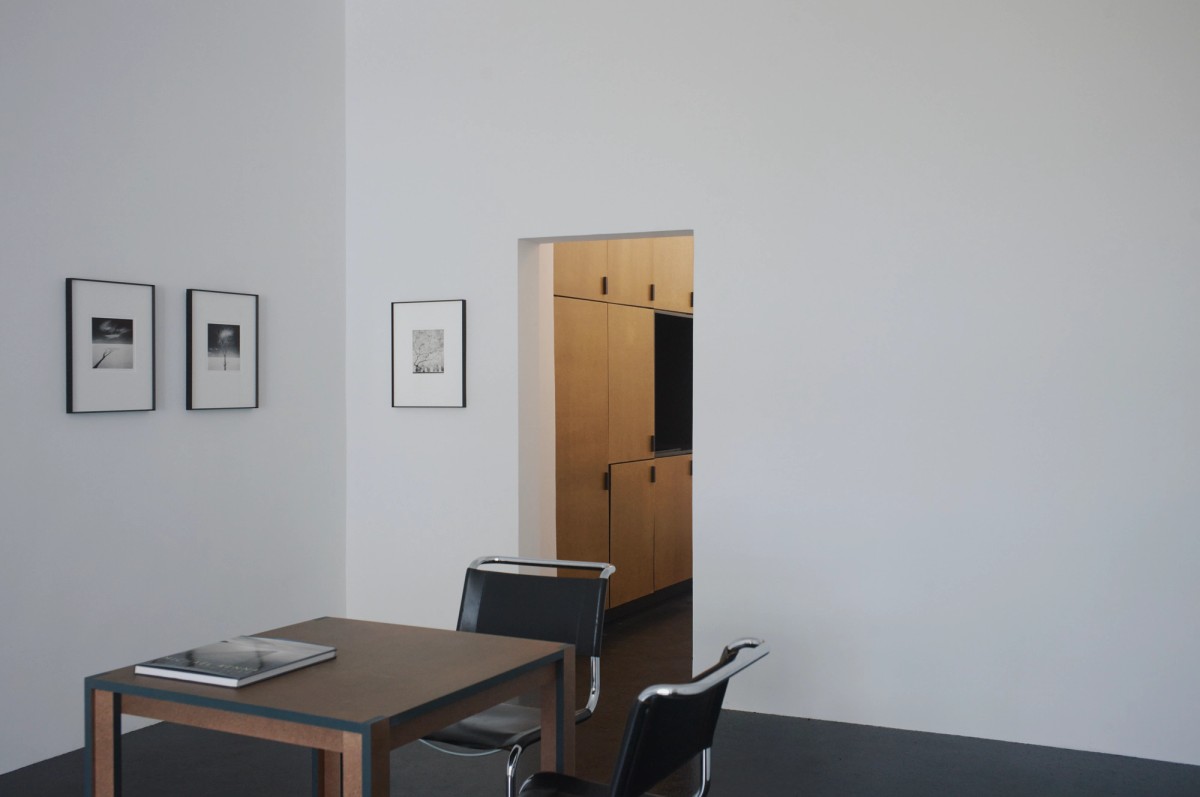 Minimalist gallery interior with a small table, two black chairs, and a photography book in the foreground. Framed black-and-white prints hang on white walls, with a warm wooden cabinet visible through a doorway, adding contrast to the clean, modern space.