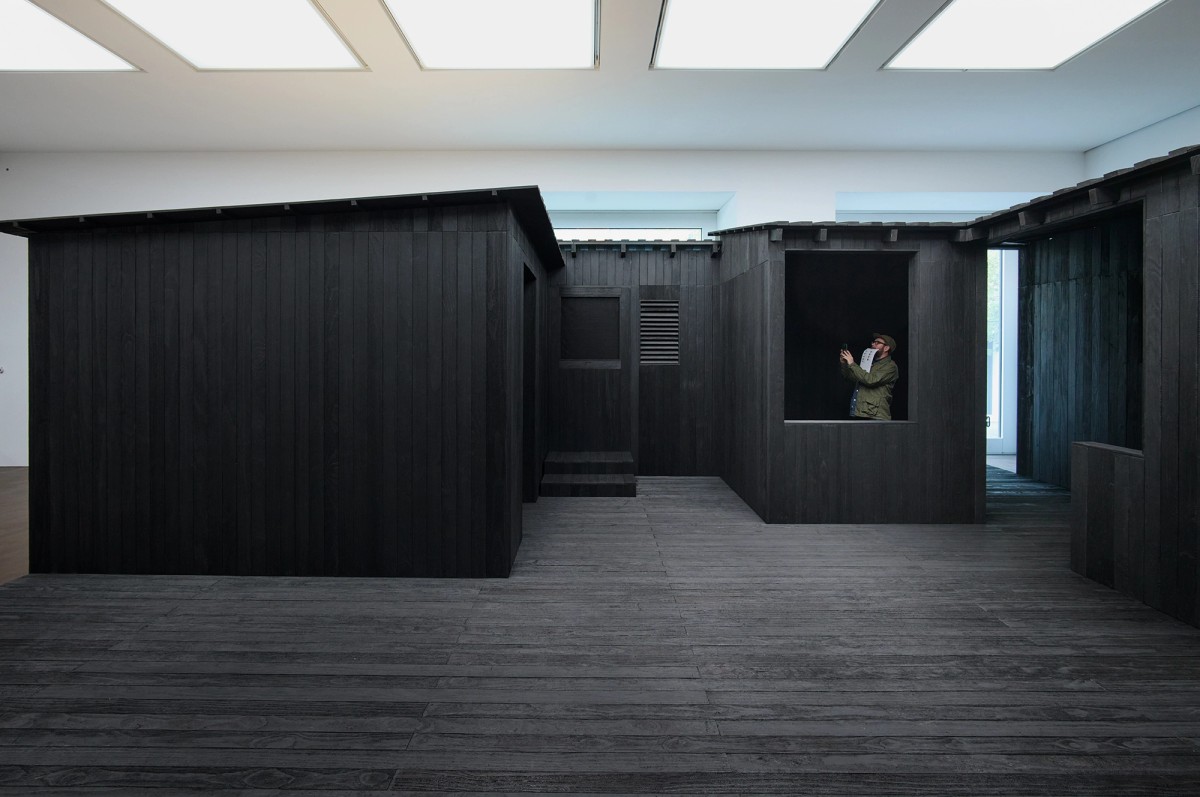 A person stands inside a large, dark wooden structure with modern, minimalist design, illuminated by overhead rectangular lights and a window in the background.