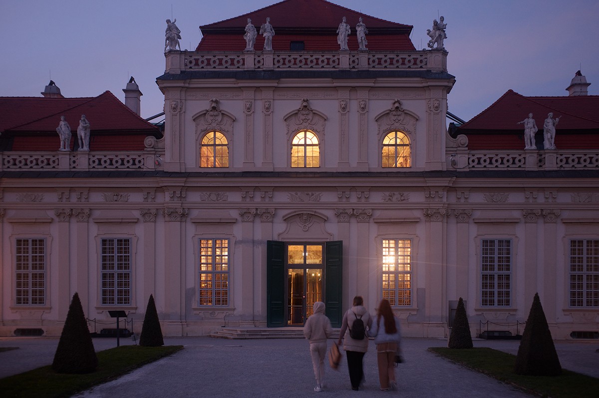 The exterior of a large building is seen from the entrance, with sunset tones coming from behind