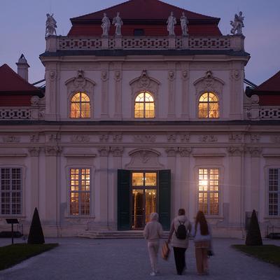 Three people walk toward the warmly lit entrance of a grand baroque building at dusk, its ornate façade adorned with sculptures and tall arched windows. The soft glow from inside contrasts with the cool twilight sky, highlighting the historic architecture and red-roofed structure.