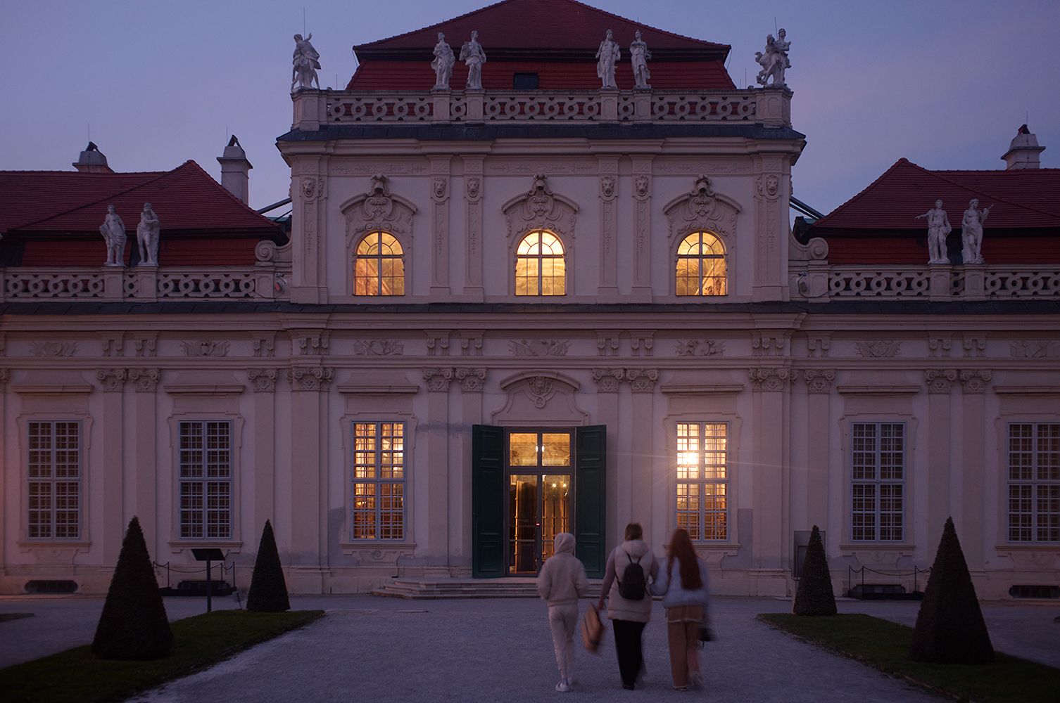 Three people walk toward the warmly lit entrance of a grand baroque building at dusk, its ornate façade adorned with sculptures and tall arched windows. The soft glow from inside contrasts with the cool twilight sky, highlighting the historic architecture and red-roofed structure.