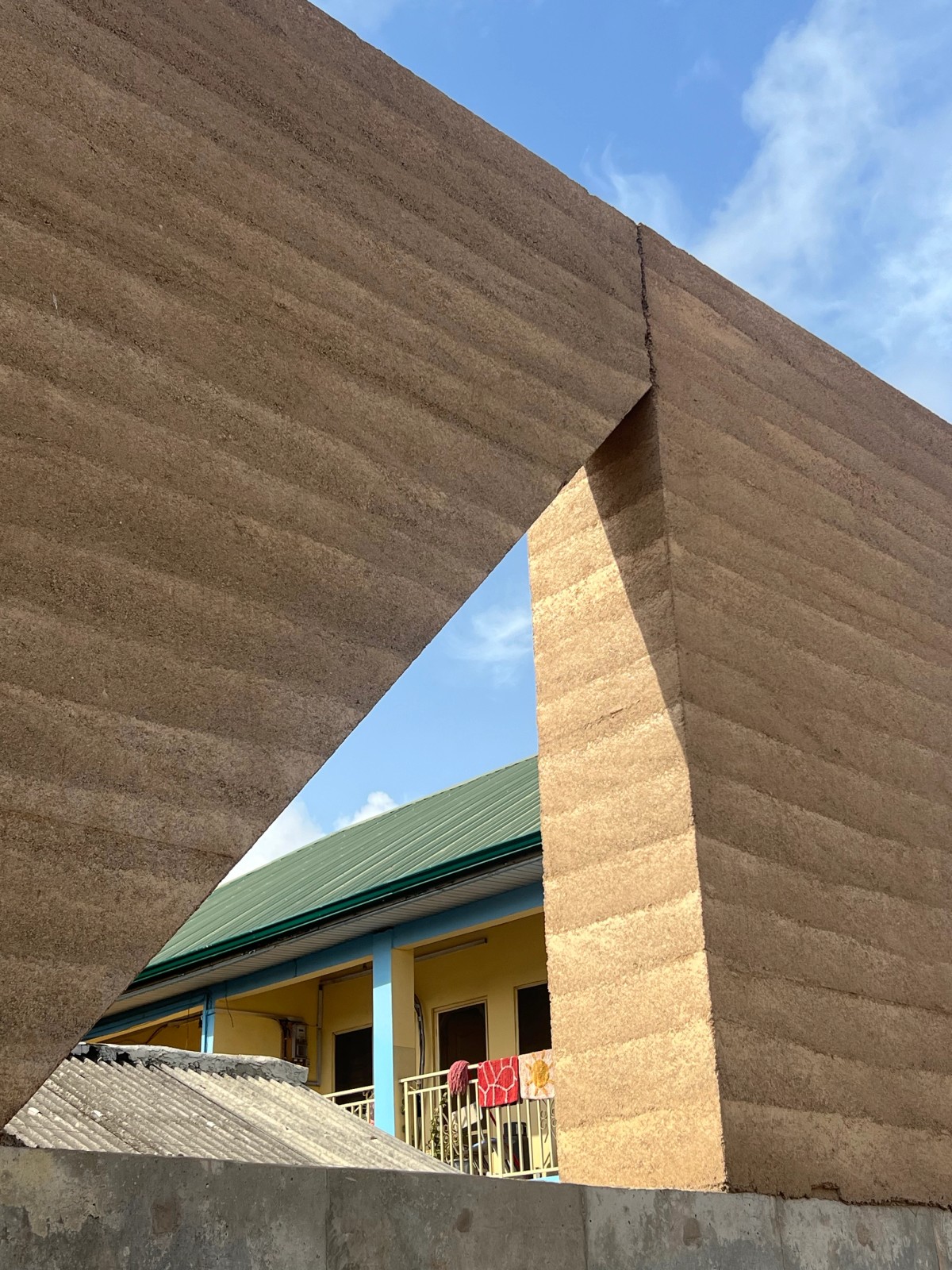 A large rammed earth wall with a triangular cut-out frames the view of a yellow building with blue columns and a green metal roof. Brightly colored towels hang over the balcony railing, adding a domestic contrast to the raw, monolithic structure in the foreground. The sky is partly cloudy with warm sunlight illuminating the scene.