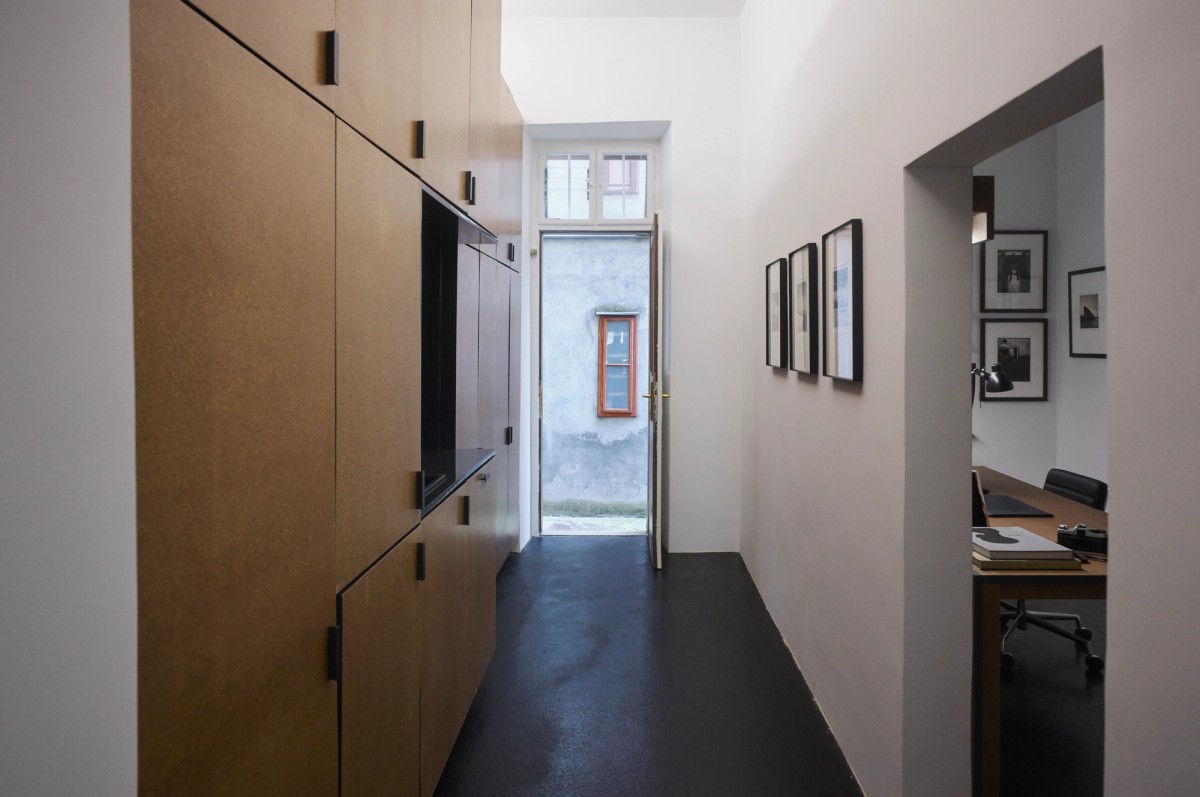 Narrow hallway with built-in wooden cabinetry on one side and framed photographs on the opposite wall, leading to an open door with a view of a textured gray exterior wall and small window. A side room reveals a minimalist office with a desk, books, and black-and-white photo prints on the walls.