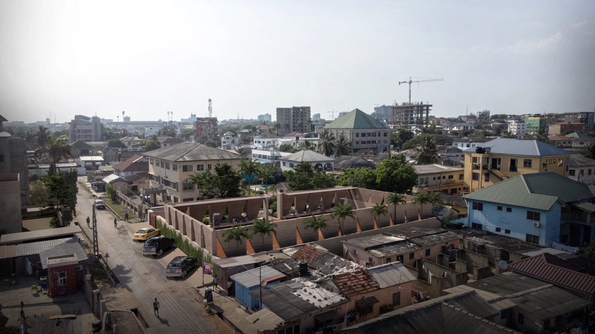 Aerial view of a mixed urban neighborhood featuring informal housing with corrugated roofs in the foreground and modern mid-rise buildings under construction in the background. A distinctive, geometric courtyard structure lined with palm trees and triangular supports stands at the center, contrasting with the surrounding architecture.