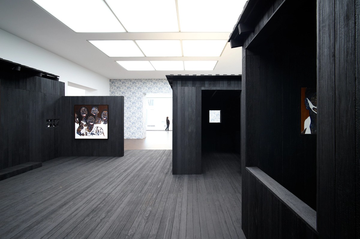 Art gallery with dark wooden structures, framed artwork on the walls, and a person walking in a brightly lit hallway in the background. The space has high ceilings and a modern, minimalist design.