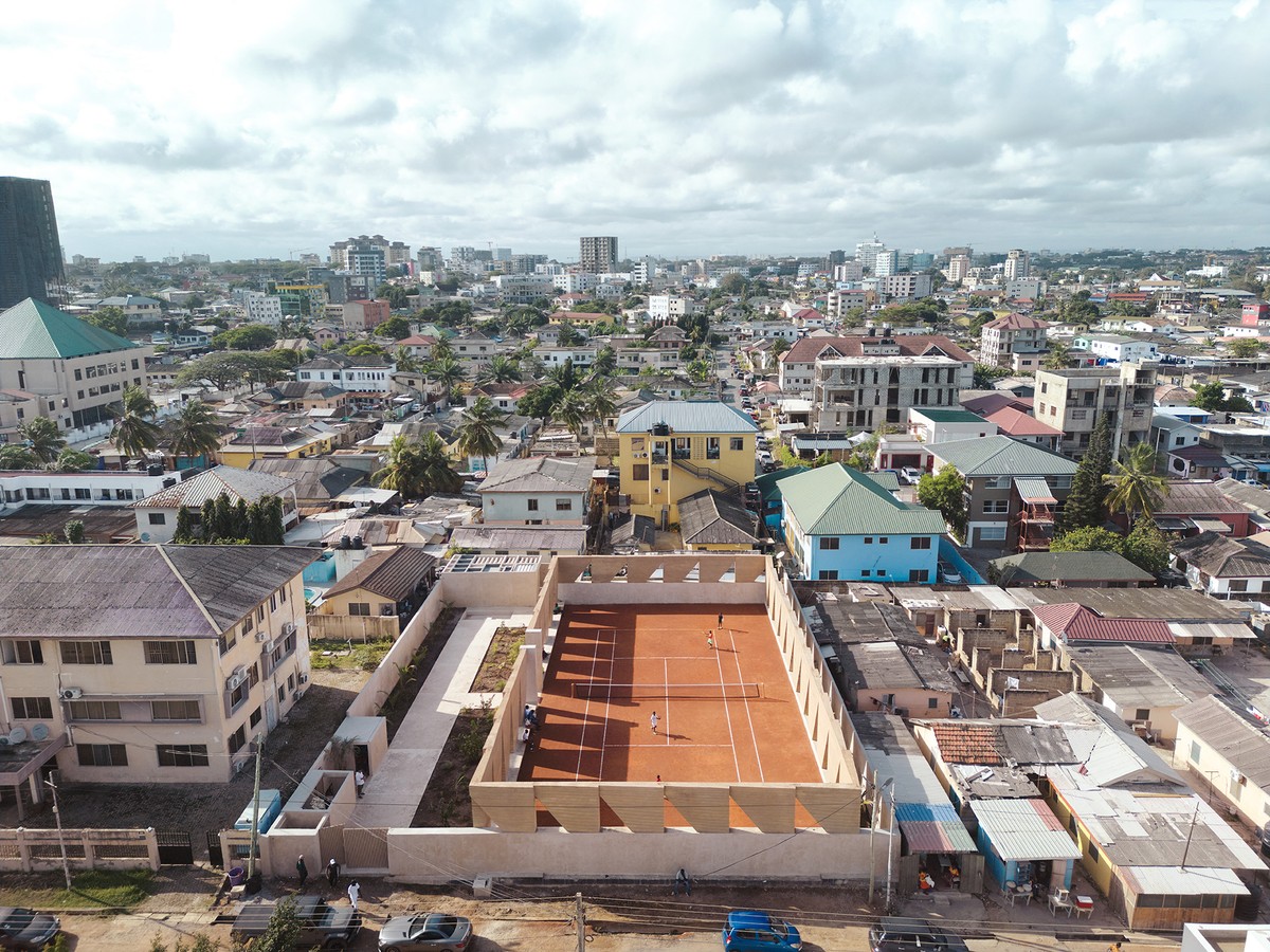 Aerial view of a mixed urban neighborhood featuring informal housing with corrugated roofs in the foreground and modern mid-rise buildings under construction in the background. A distinctive, geometric courtyard structure lined with palm trees and triangular supports stands at the center, contrasting with the surrounding architecture.
