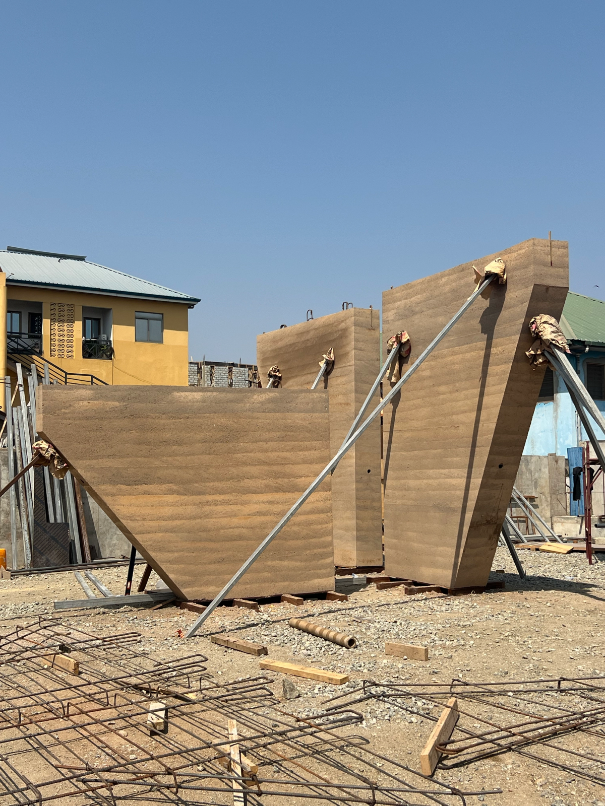 A wooden structure being propped up and suspended next to a earthy toned building and in front of a deep blue sky.