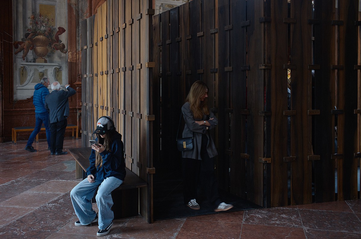 Inside an ornate, historic interior with marble floors and painted walls, people interact with a large, dark wooden installation composed of vertical slats. One person sits on a bench using their phone, while others explore or observe the structure, blending contemporary design with a classical setting.