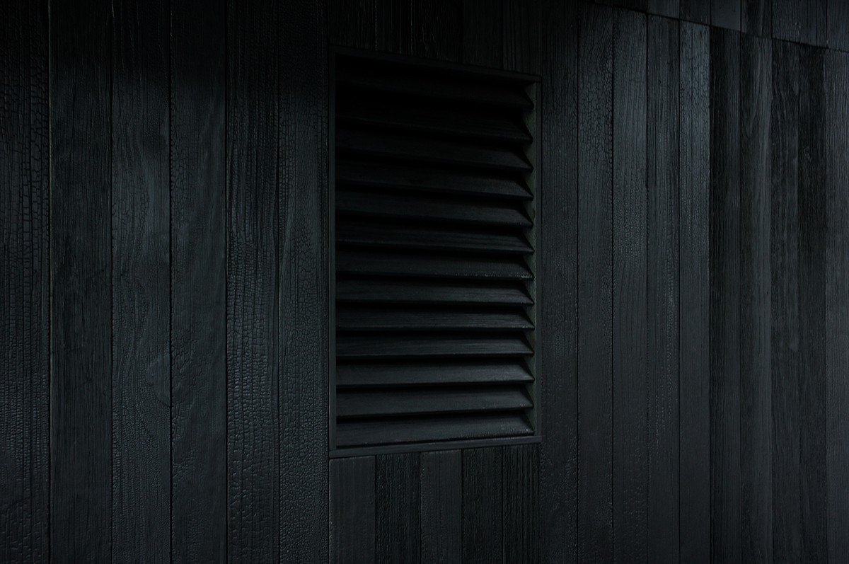 A dark, textured wooden wall with vertical planks features a rectangular black vent with horizontal slats near the center. The overall lighting is dim, creating a moody and shadowy atmosphere.