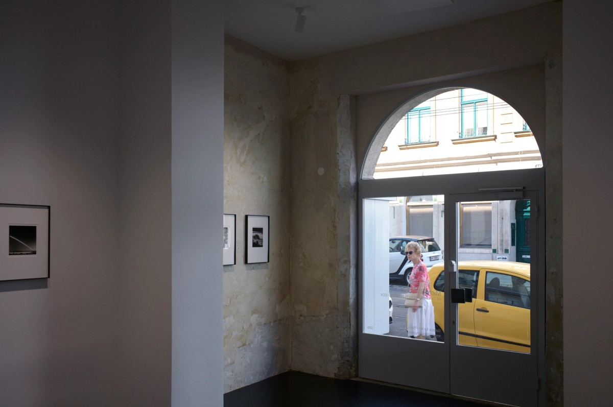 Interior of a minimalist gallery with white and weathered walls displaying black-and-white photographs. Sunlight streams through an arched window above a glass door, where a woman in a red-striped shirt passes by a parked yellow car, blending street life with the quiet exhibition space.