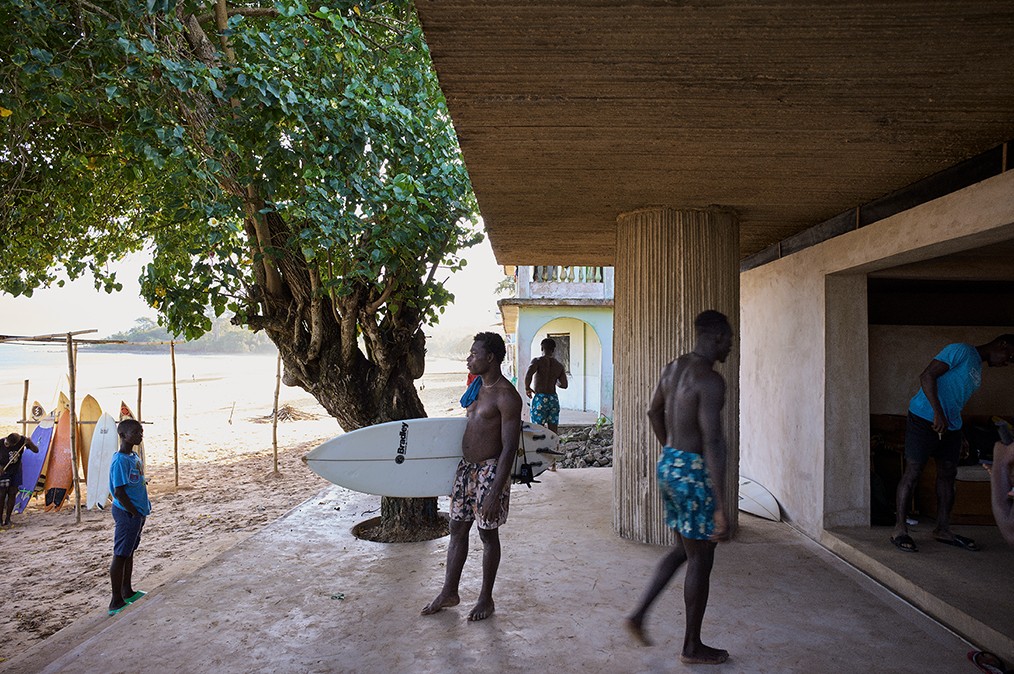 A figure stands between a building and beach, under a roof and tree, with a surfboard