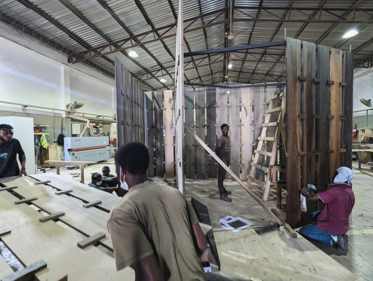 Inside a spacious workshop with a corrugated metal roof, several workers assemble a large, circular wooden structure composed of dark vertical panels. Some workers are actively sanding or positioning pieces, while others handle tools or observe the construction