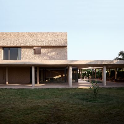 Modern residence with a textured beige facade and flat roof, featuring a minimalist colonnade that extends into a covered patio area. Floor-to-ceiling glass walls reveal open interior spaces, while a manicured lawn and tropical plants enhance the tranquil, sunlit outdoor setting.