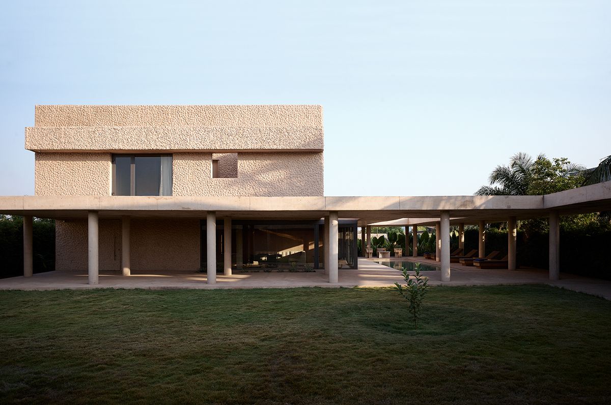 Modern residence with a textured beige facade and flat roof, featuring a minimalist colonnade that extends into a covered patio area. Floor-to-ceiling glass walls reveal open interior spaces, while a manicured lawn and tropical plants enhance the tranquil, sunlit outdoor setting.
