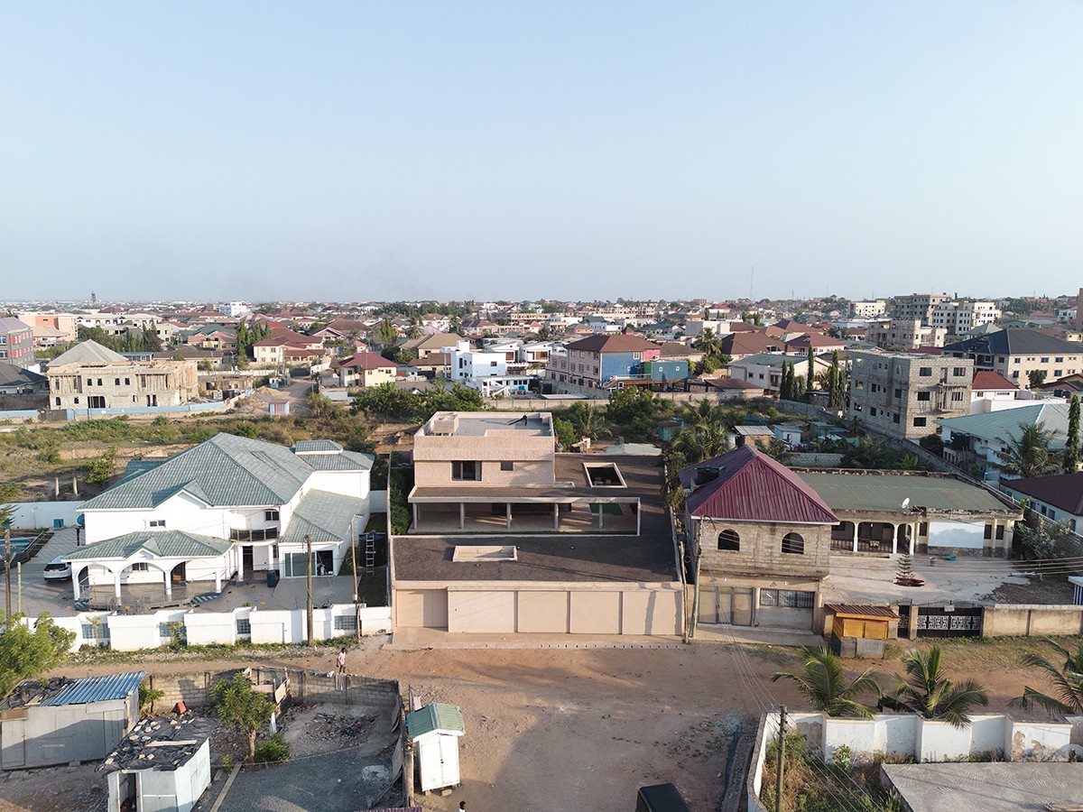 Aerial view of a residential neighborhood featuring a mix of finished and under-construction homes with various roof styles and colors. In the foreground, a modern, boxy structure with a textured facade contrasts with more traditional buildings, set against a dense urban landscape that stretches to the horizon under a clear sky.