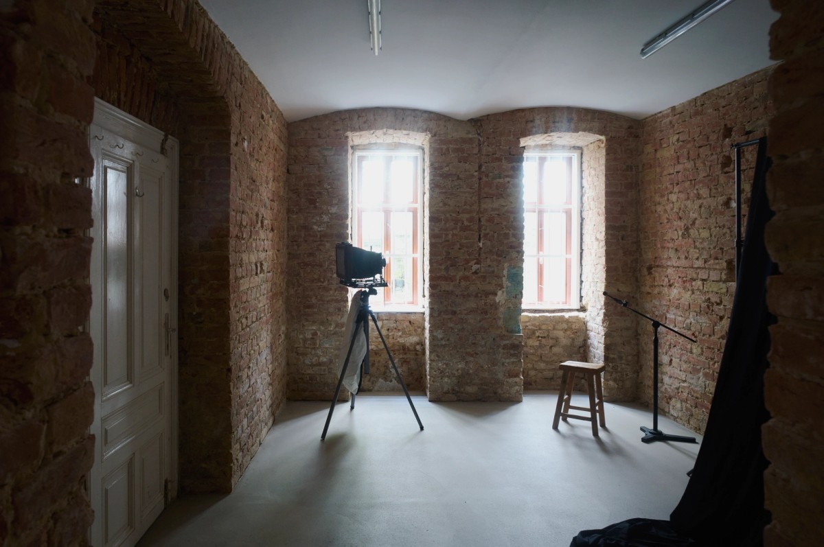A dimly lit room with exposed brick walls and arched windows contains a large-format camera on a tripod, a wooden stool, and a microphone stand. Natural light from the windows highlights the texture of the brick, creating a quiet, atmospheric setting that feels both historic and suited for portrait photography or recording