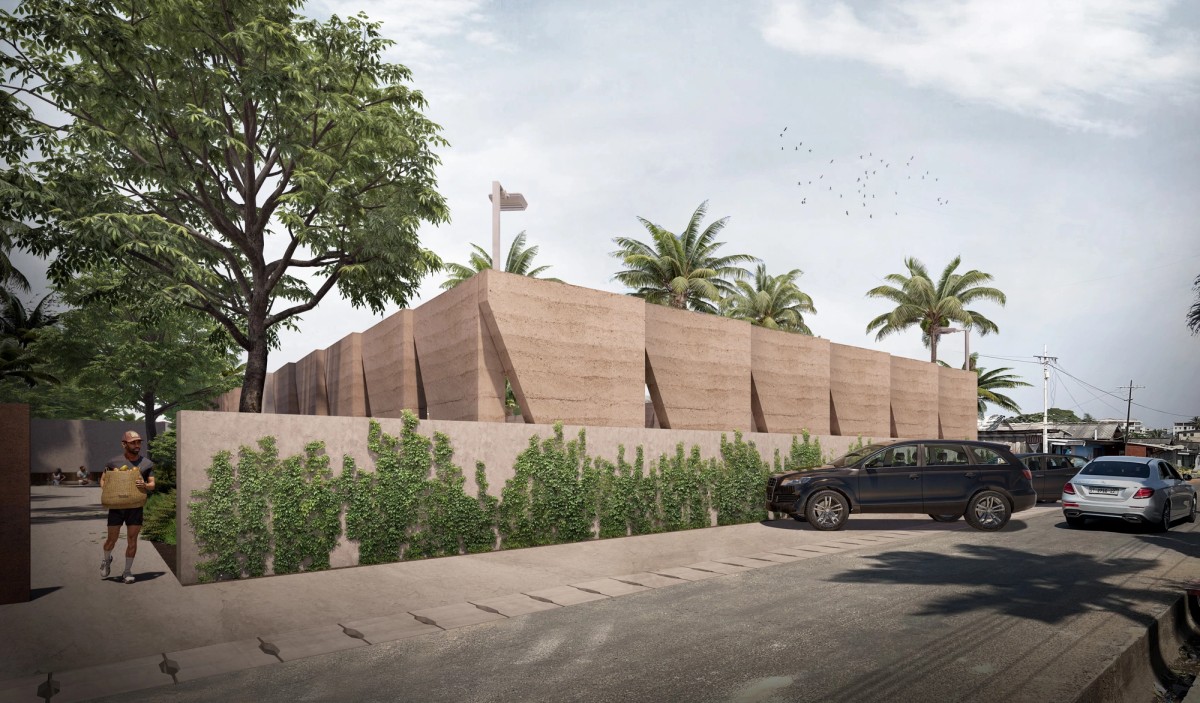 A street-level view of a contemporary building with geometric rammed earth walls, partially obscured by a low wall covered in creeping green vines. Palm trees and other lush vegetation surround the site, giving it a tropical feel. Parked cars line the road, while a man carrying a woven basket walks toward the entrance. The angular structure contrasts with the surrounding traditional neighborhood, blending natural materials with a minimalist design. A flock of birds is visible in the sky above.