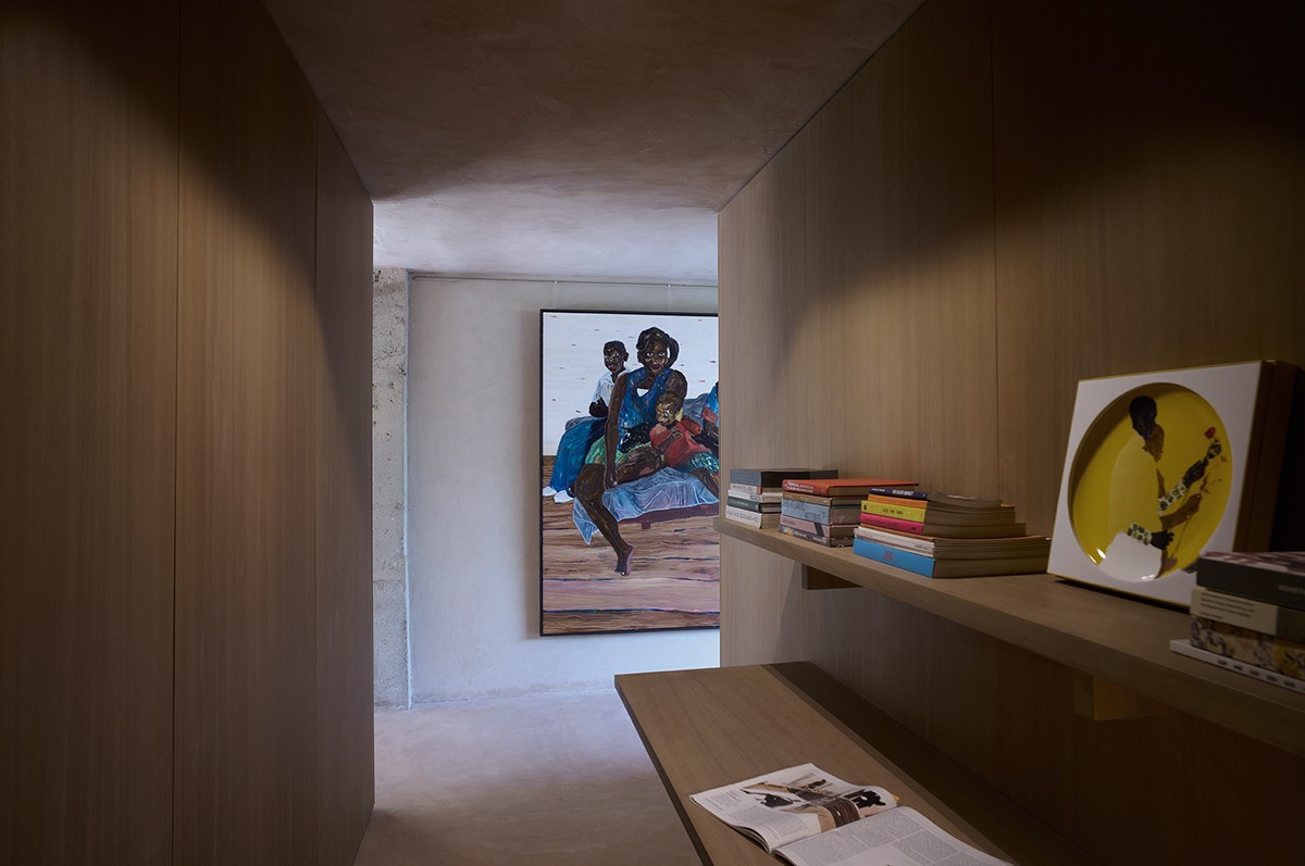 A narrow, softly lit hallway lined with wooden paneling features built-in shelving filled with books and art objects. At the end of the corridor, a large, colorful painting of a family seated together adds vibrancy and warmth to the otherwise minimalist space.