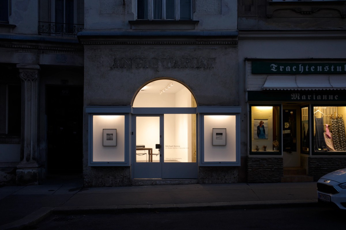 Evening street view of a softly illuminated photography gallery with a central arched doorway flanked by two framed black-and-white prints in window displays. The gallery’s clean, minimal interior contrasts with the worn façade labeled “ANTIQUARIAT,” situated between darker storefronts, including a neighboring traditional clothing boutique.
