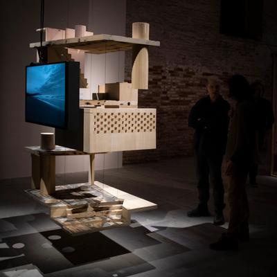 Wooden architectural sculpture with suspended monitor showing ocean scene, exhibited in a dimly lit gallery.