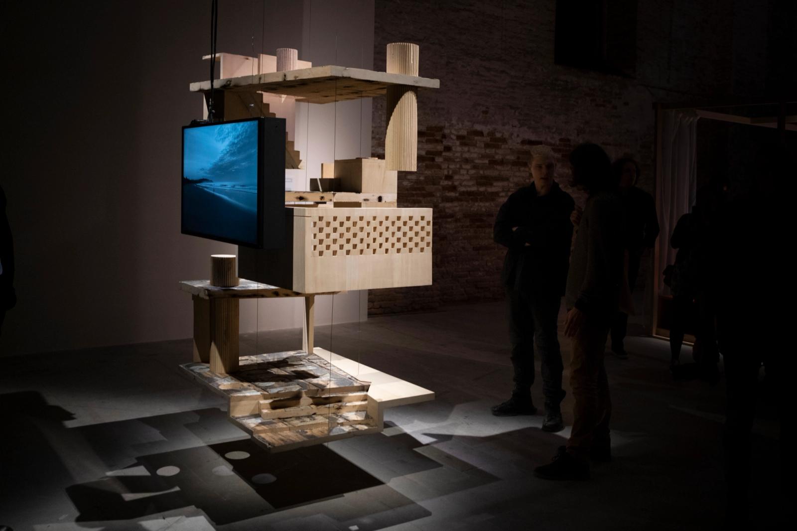 Wooden architectural sculpture with suspended monitor showing ocean scene, exhibited in a dimly lit gallery.