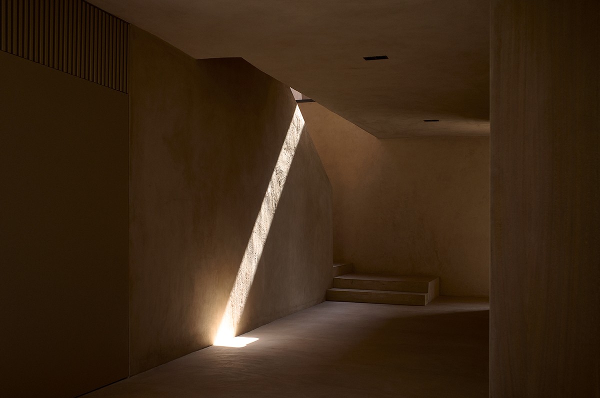 A narrow beam of natural light cuts through a dark, minimal interior with textured beige walls and clean architectural lines. The space features subtle steps and recessed ceiling lights, creating a quiet, contemplative atmosphere defined by shadow and simplicity.