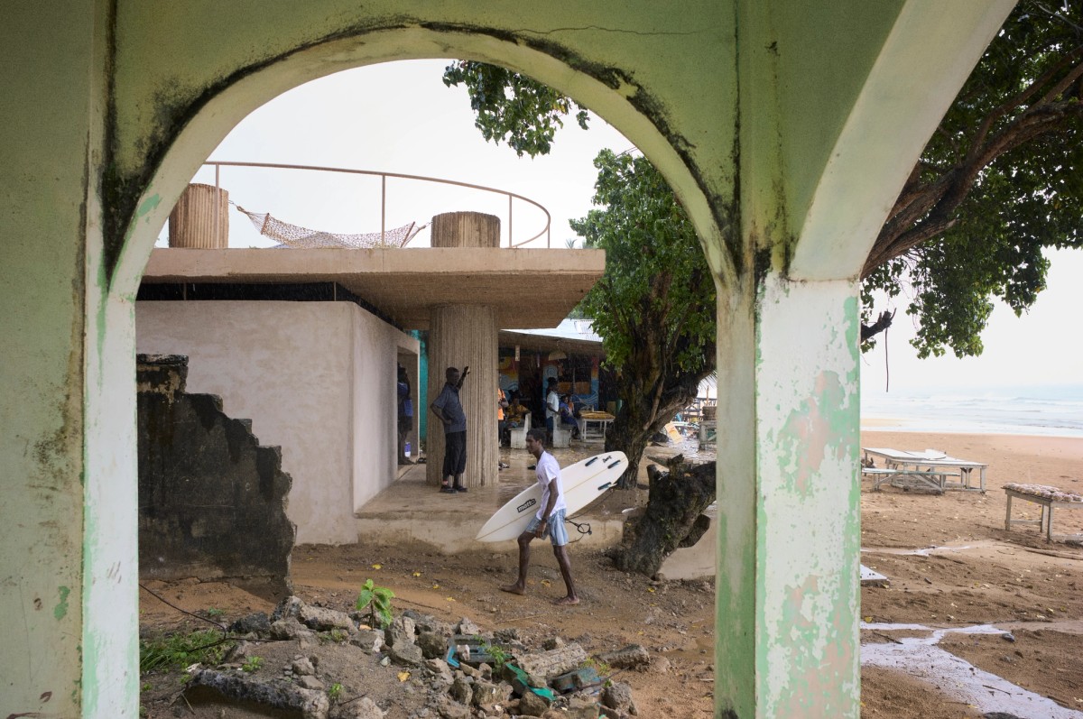 Framed by a weathered green archway, the image shows a beachside structure with exposed concrete columns and a gathering of people beneath a large tree. A man carrying a surfboard walks across the sandy ground, evoking a laid-back, coastal atmosphere near the ocean.