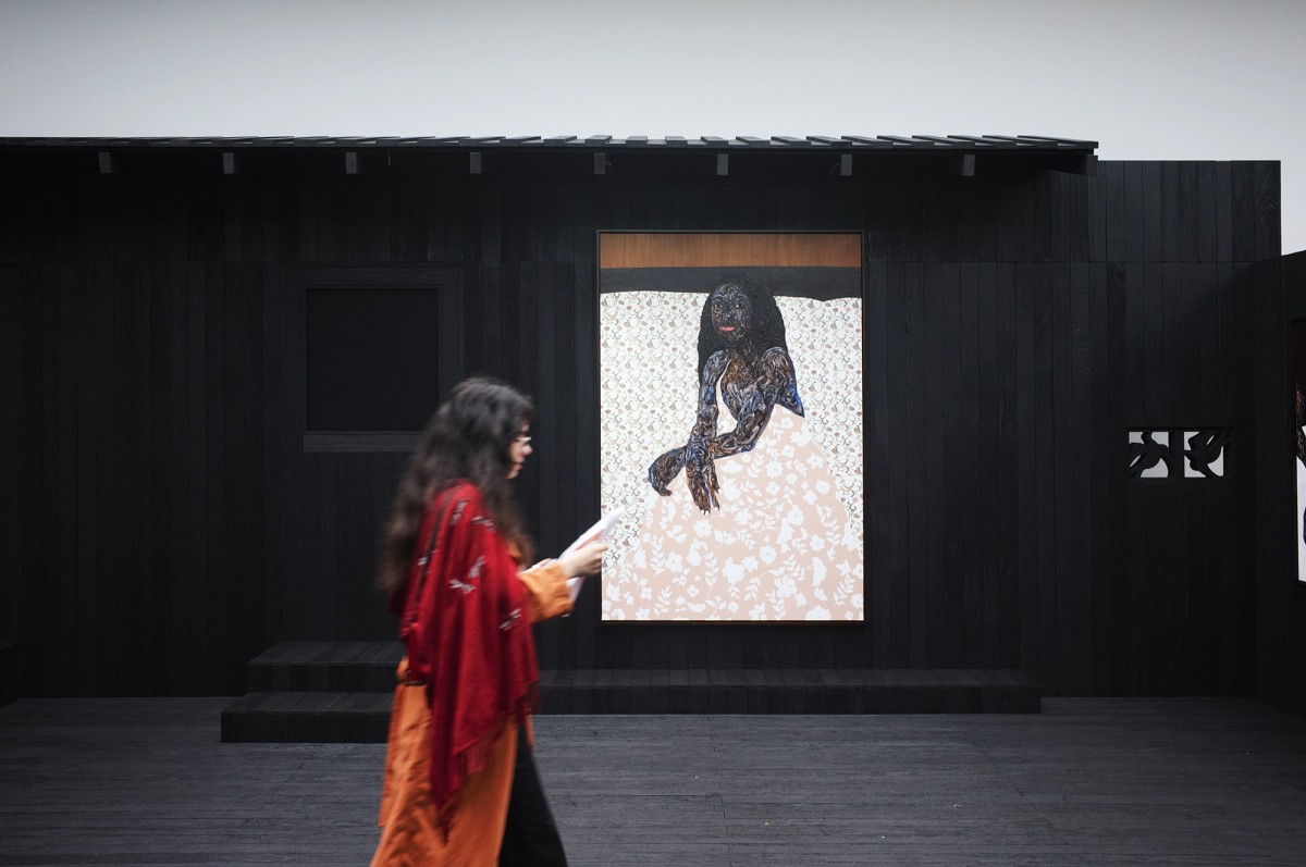 A person in an orange and red outfit walks past a dark wooden structure displaying a large artwork of a seated woman with dark skin and textured hair, set against a light floral background.
