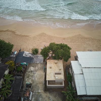 Framed by a weathered green archway, the image shows a beachside structure with exposed concrete columns and a gathering of people beneath a large tree. A man carrying a surfboard walks across the sandy ground, evoking a laid-back, coastal atmosphere near the ocean.