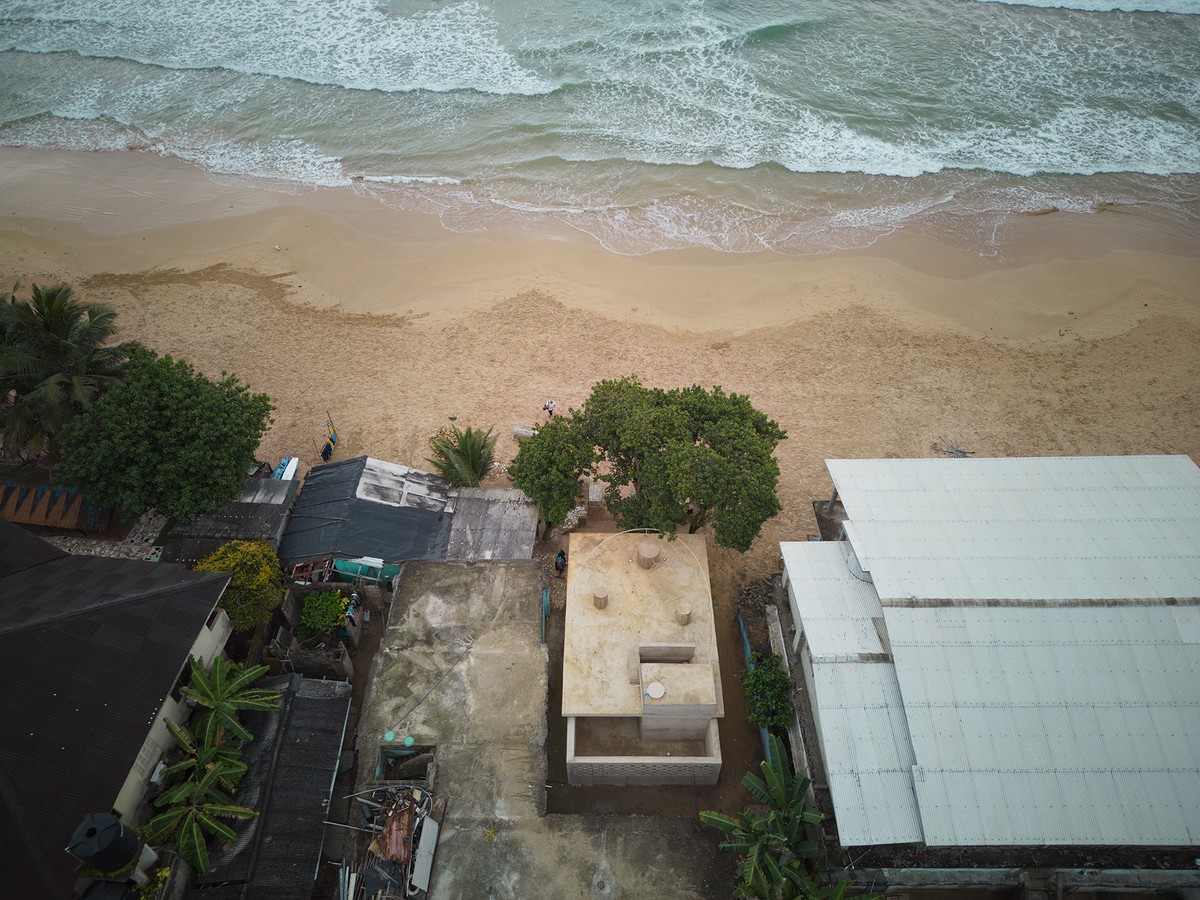 Aerial view of a coastal area showing ocean waves hitting a sandy beach. Just inland, several small buildings with metal and concrete roofs are surrounded by trees and vegetation.