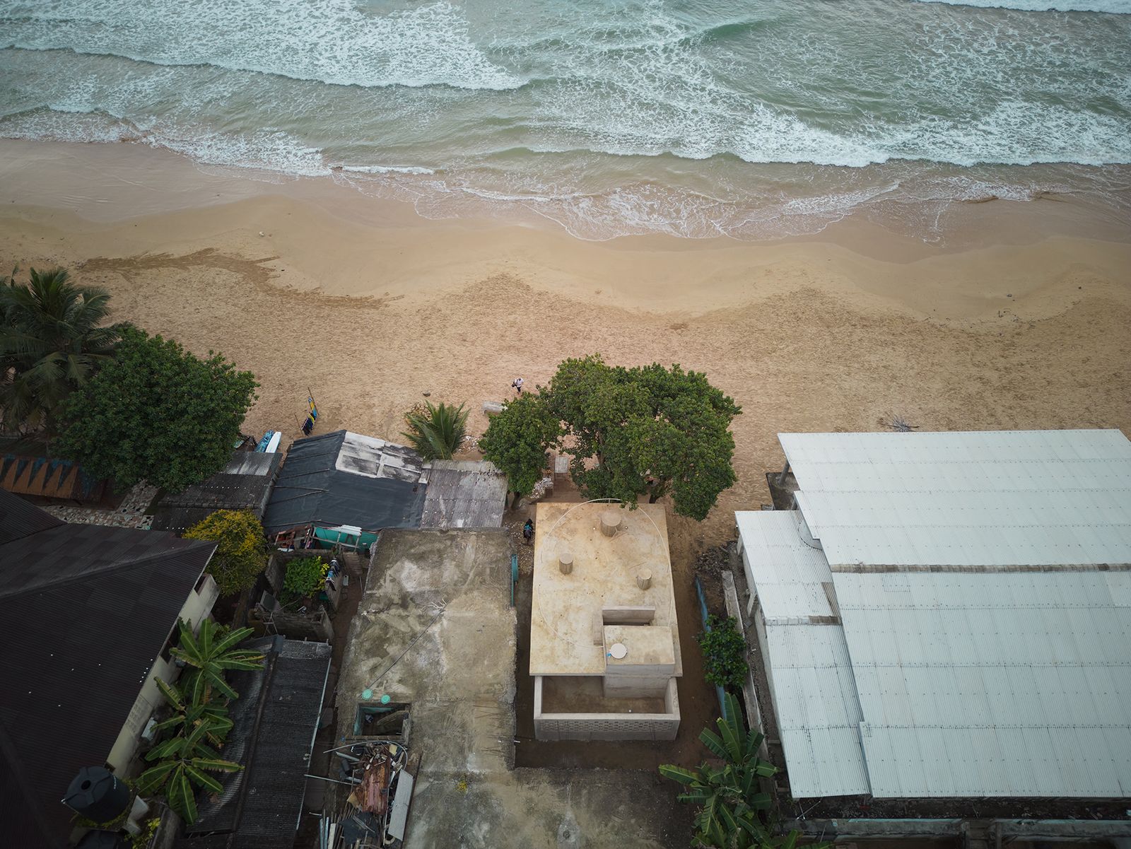 Framed by a weathered green archway, the image shows a beachside structure with exposed concrete columns and a gathering of people beneath a large tree. A man carrying a surfboard walks across the sandy ground, evoking a laid-back, coastal atmosphere near the ocean.