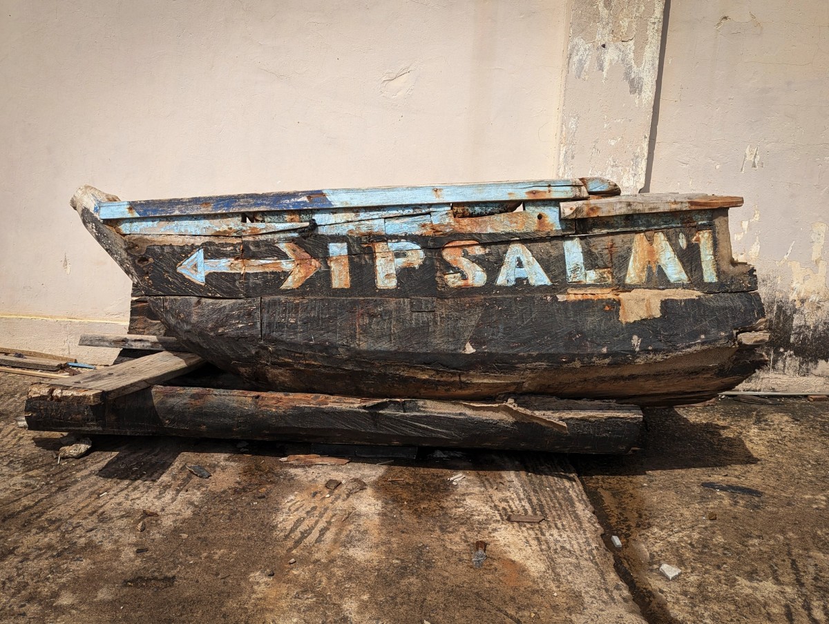 Weathered wooden boat fragment with faded blue text reading “PSALM” and directional arrows, leaning against a sunlit wall.