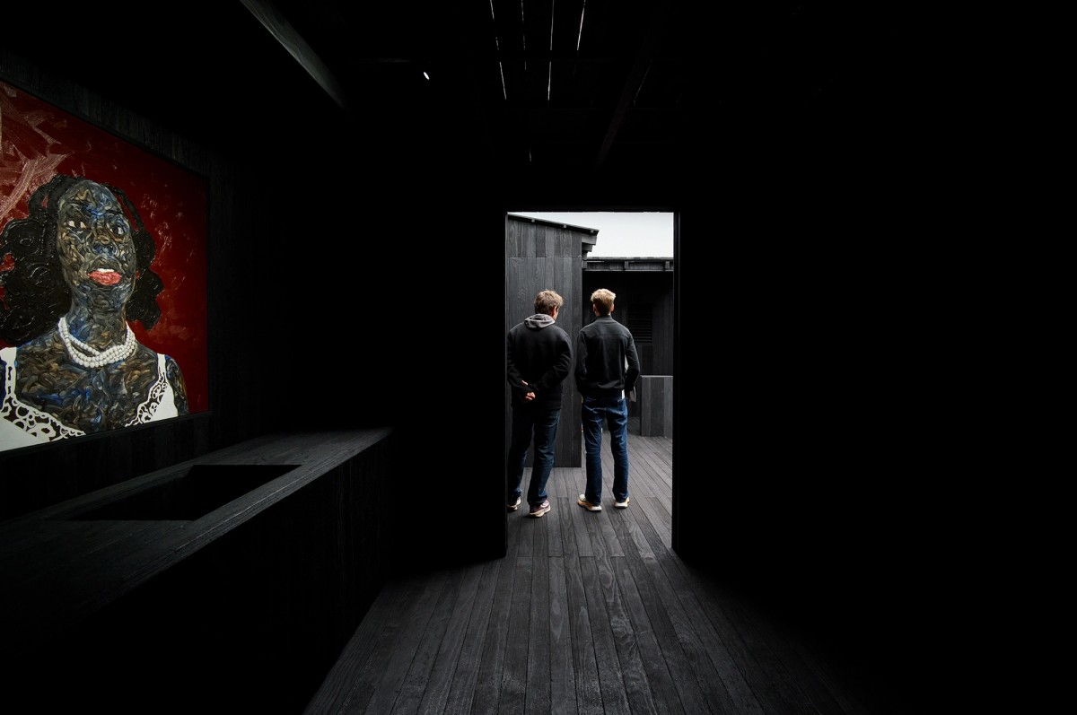 Two people stand in a dark, minimalist room and look outside through an open doorway. A colorful portrait of a woman with a red background hangs on the left wall. The space is dimly lit, creating a dramatic contrast.
