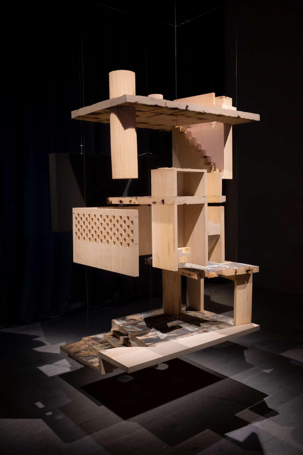 Full view of a layered, suspended wooden sculpture resembling a fragmented building, dramatically lit from above.
