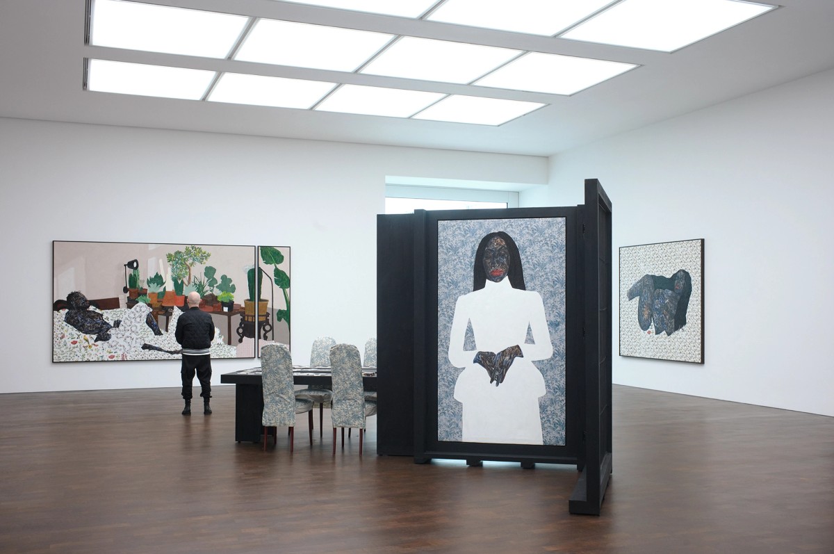 Art gallery with dark wooden structures, framed artwork on the walls, and a person walking in a brightly lit hallway in the background. The space has high ceilings and a modern, minimalist design.