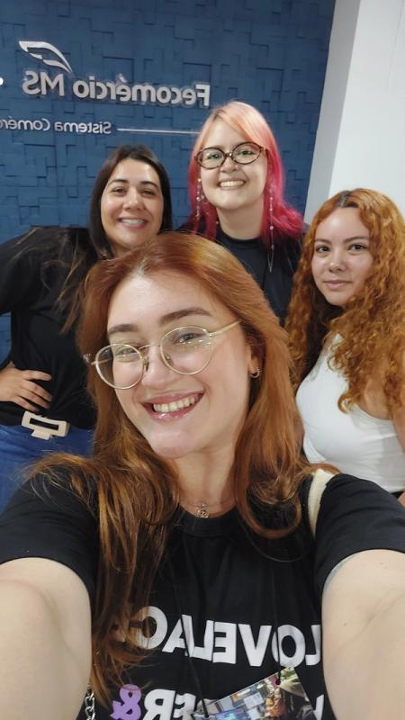 Quatro mulheres sorrindo em uma selfie, em ambiente interno, com logo ao fundo e clima descontraído.