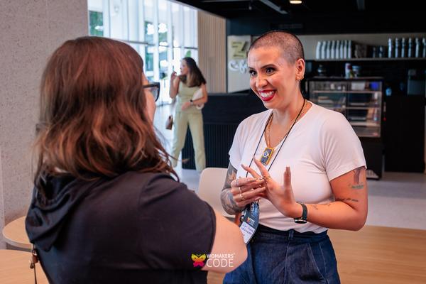 Duas mulheres conversando em um evento; uma delas gesticula enquanto fala, ambas usam crachá da WoMakersCode