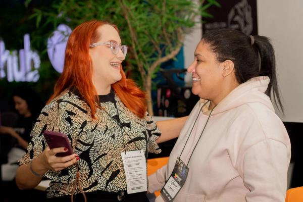 Duas mulheres sorrindo e conversando em um evento, usando crachás da WoMakersCode, com fundo desfocado